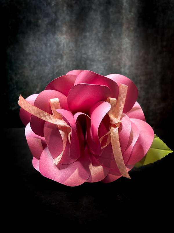 Primo piano frontale di un bouquet portafedi artigianale a forma di grande rosa color rosa ciclamino con sfumature perlate, foglia verde laterale e due nastri color pesca inseriti tra i petali per legare le fedi nuziali, su sfondo scuro.