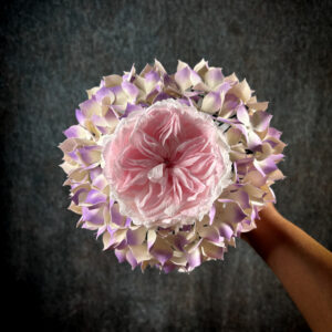 Vista dall'alto di un bouquet sposa con ortensie di carta rigida sfumata lilla e crema che circondano una rosa inglese rosa confetto.