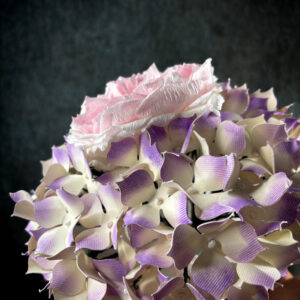 Vista dall'alto di un bouquet sposa con ortensie di carta rigida sfumata lilla e crema che circondano una rosa inglese rosa confetto.
